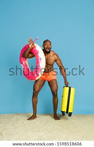 Shocked. Happy young man resting with beach ring as a donut and bag on blue studio background. Concept of human emotions, facial expression, summer holidays or weekend. Chill, summertime, sea, ocean.