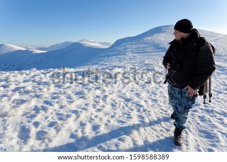 Young man photographer in winter clothing standing and making photo with camera in sunlight with white snow background. Travelling and making photos of winter nature concept