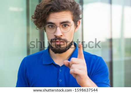 Serious concerned guy pointing index finger up. Handsome bearded young man in blue casual t-shirt standing at outdoor glass wall. Warning concept