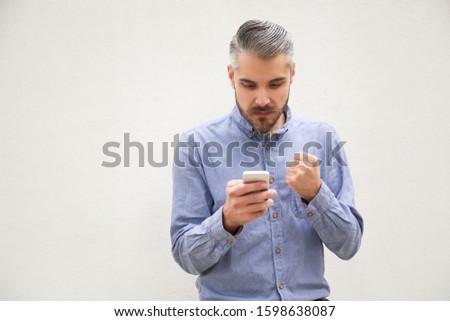 Serious stressed man clenching fist, reading message on smartphone. Grey haired young man in blue casual shirt posing isolated over white background. Wireless internet connection concept