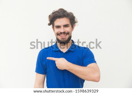 Cheerful positive customer pointing finger away at copy space. Handsome bearded young man in blue casual t-shirt posing isolated over white background. Advertising concept