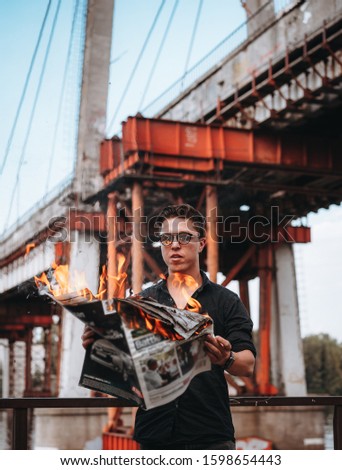 Guy reads a burning newspaper, in the background a bridge
