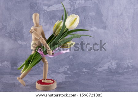 Wooden dummy holding fresh white tulips bouquet near red decorative hearts laying on table. Stepping man figure on grey background. Copy space for text