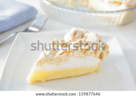 a slice of home made lemon meringue pie on a plate, with the pie dish and a fork and napkin in the background