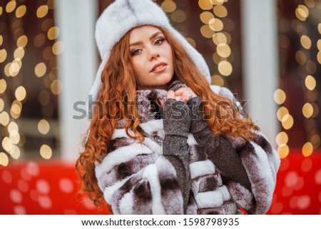 Outdoor portrait of young beautiful redhead happy smiling girl is wearing fur warm hat. Festive Christmas lights on background.