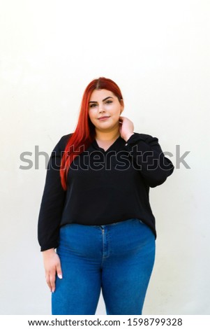 Plus size model with long red hair posing on a street wall. Caucasian ethnicity woman dressed in black shirt and blue pants. Concept of body positivity. Royalty-Free Stock Photo #1598799328