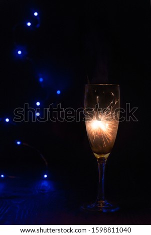 Sparkler inside a glass of champagne, which stands on a wooden table in the dark, in the background are blue bokeh lights.