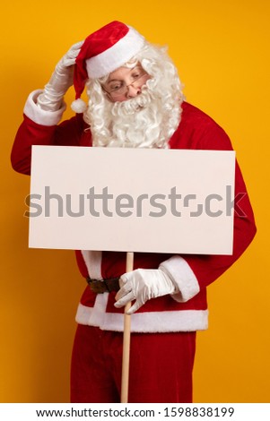 Santa Claus holds in his hands large white billboards on a stick for recording ads and posing on a yellow background