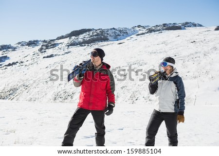 Smiling couple carrying ski boards on shoulders while walking on snow covered landscape