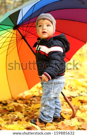 Happy playful child outdoors on autumn leaves. Little boy in the autumn park with rainbow umbrella. Background of autumn trees.Baby boy walking in the park