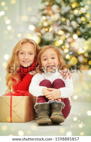 Merry Christmas and Happy New Year! Happy sisters are sitting next to a box with a Christmas gift at home beautifully decorated for holidays. Christmas by a beautiful Christmas tree. 