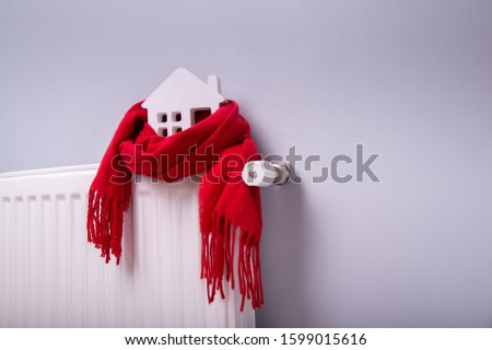 White house model covered in warm red scarf against gray wall. Model of house on white radiator.