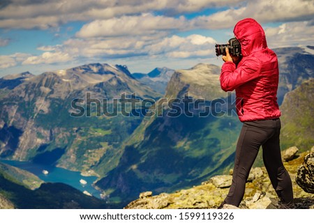 Tourism vacation and travel. Female tourist taking photo with camera, enjoying Geiranger fjord and mountains landscape from Dalsnibba Plateau viewpoint at windy day, Norway .