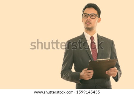 Studio shot of young Asian businessman holding clipboard while thinking