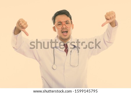 Studio shot of young angry Asian man doctor giving thumbs down