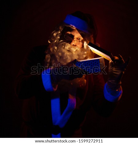 Santa Claus holds a gift box in his hands, looks into it and poses on a dark red background