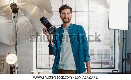 In the Photo Studio with Professional Equipment: Portrait of the Handsome Photographer Holding State of the Art Camera Ready to Take Pictures with Softboxes Lighting in Background