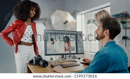 In Photo Studio Fashion Magazine Designer and Beautiful Black Cover Girl talk to a Professional Photographer who Uses Desktop Computer to Retouch Photographs in an Image Editing Software