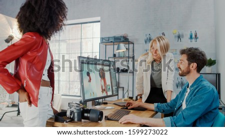 In Photo Studio Fashion Magazine Designer and Beautiful Black Cover Girl talk to a Professional Photographer who Uses Desktop Computer to Retouch Photographs in an Image Editing Software