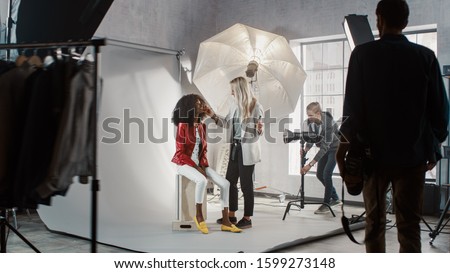 Preparations on a Backstage of the Photo Shoot: Make-up Artist Applies Makeup on Beautiful Black Model, Assistant Adjusts the Lightning Equipment. Fashion Magazine Studio Photoshoot