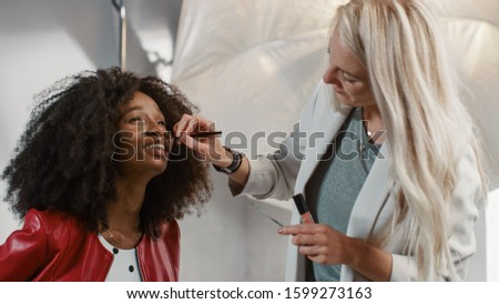 Backstage of the Photo Shoot: Make-up Artist Applies lipstick Makeup on Beautiful Black Model with Lush Curly Hair. Fashion Magazine Studio Photoshoot Preparations. Professional Equipment Studio