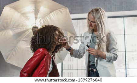 Backstage of the Photo Shoot: Make-up Artist Applies Makeup on Beautiful Black Model. In the Background Studio with Modern Photo Equipment. Fashion Magazine Studio Photoshoot