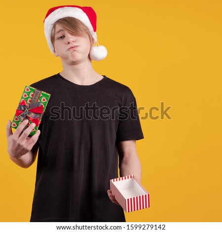 Young guy in a red santa hat holds a gift box and money in his hands and poses on a yellow background