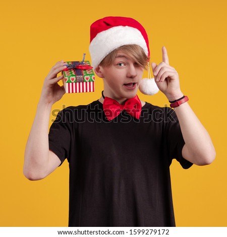 Young guy in a red santa hat holds a gift box and money in his hands and poses on a yellow background