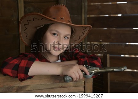 young girl cowboy in a red checkered shirt on the background of a wooden wall