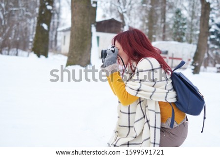 Young redhead smiling female in warm clothes with retro film camera shooting a photo on the background of winter forest, snow