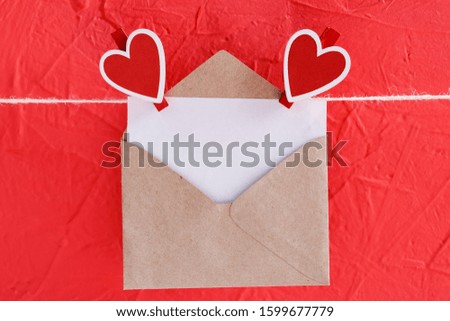 empty sheet of paper in envelope hangs on a rope attached with heart-shaped clothespins on a red background. concept of Valentine's day, anniversary, mother's day or birthday greeting, copy space