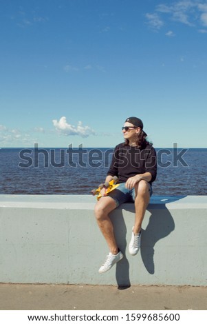 A young man with long hair in a baseball cap, denim shorts, sweatshirt and black glasses is sitting with a yellow skate on the city's quay. Copy space right.
