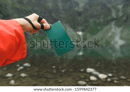 Male hand holding a green blank card on a mountain lake background. Green flyer in the hands of man on the background of untouched nature, mountains and lake. Copy space