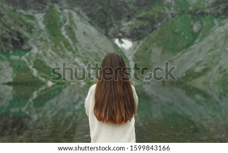 Brunette girl in a sweater stands on a mountain background with a lake, the view from the back. Hipster girl's back near mountain lake. Background. Hipster photo on nature. Copy space