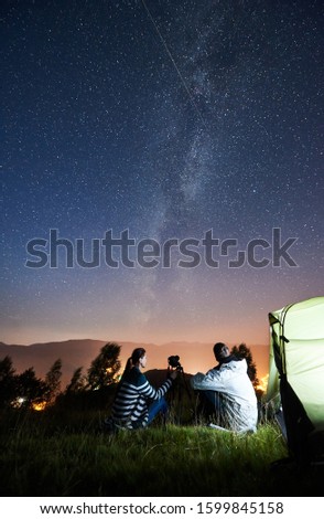 Young couple hikers resting beside camp and tourist tent with camera on tripod, making photo of amazing night sky full of stars and Milky way. On the background starry sky, mountains and luminous town