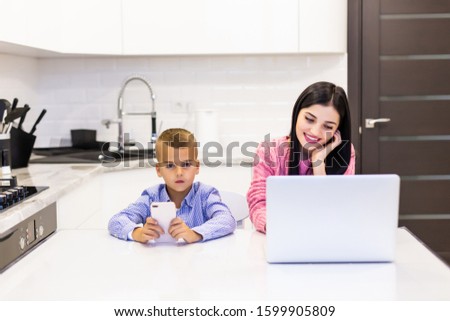 Young woman using a laptop computer for work at home while her son playing games in the phone.
