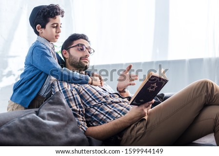 jewish father pointing with hand at tanakh and talking with son in apartment  Royalty-Free Stock Photo #1599944149
