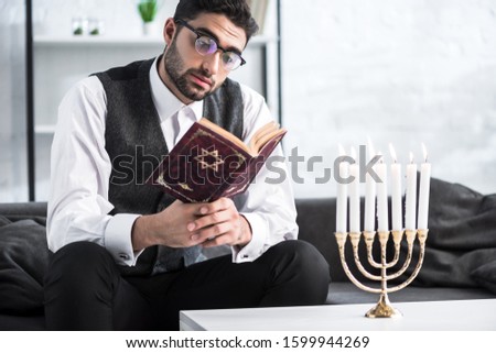handsome jewish man in glasses reading tanakh in apartment  Royalty-Free Stock Photo #1599944269