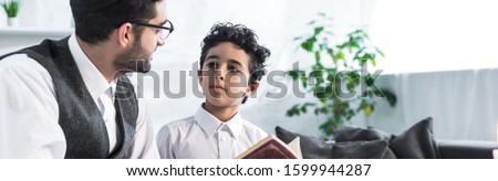 panoramic shot of jewish father and son talking and holding tanakh in apartment  Royalty-Free Stock Photo #1599944287
