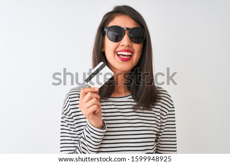 Young chinese woman wearing sunglasses holding credit card over isolated white background with a happy face standing and smiling with a confident smile showing teeth