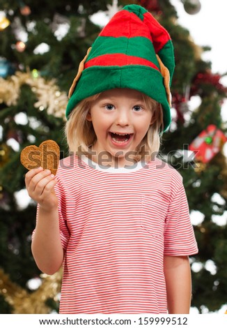 A cute excited boy dressed as Santa's helper is holding gingerbread cookie in front of a Christmas tree.