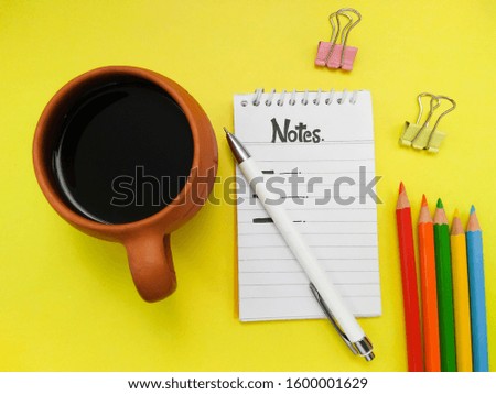 Top view of white spiral notepad diary, pen, paper clips and colorful sketch pencils with coffee mug on yellow colored background.