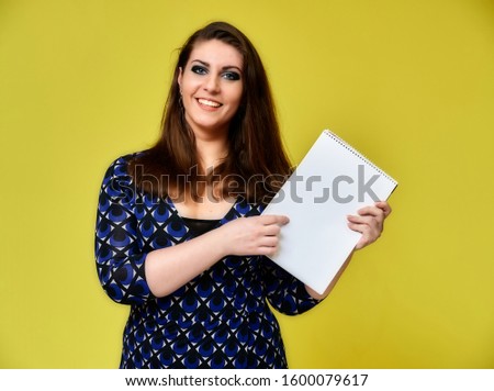 Portrait of a pretty brunette woman with a folder in her hands with a beautiful hairstyle and with excellent makeup in a dark blue blouse on a yellow background. Stands with a smile.