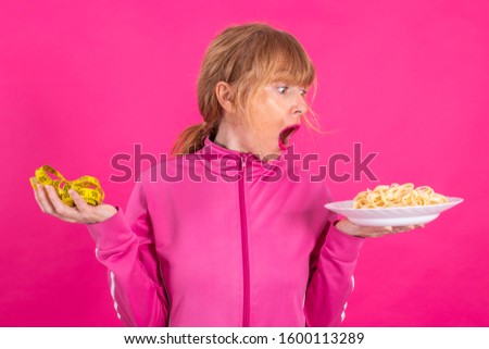 woman with stress expression looking at measuring tape and plate of pasta or hydrates with sportswear isolated on pink background