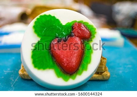 A round white piece of handmade soap with the image of two red strawberries with green leaves stands on a blue substrate.