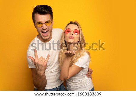 Like rock stars. A beautiful excited couple in bright sunglasses are hugging and smiling while showing the sign of the horns and blowing an air kiss to the camera.