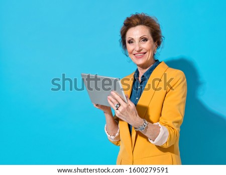 Smiling elderly woman using digital tablet. Photo of business lady in yellow blazer on blue background. Emotions and pleasant feelings concept.