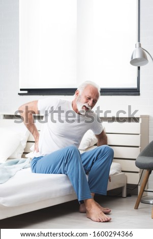 Senior man with white hair and beard alone sitting on bed and suffering from low back pain. Old age, health problem and people concept. Copy space image