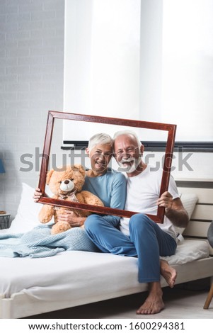 Senior couple in their pajamas on the bed with their hands hold a wooden frame while the photographer paints them. Copy space image