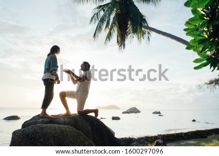 The guy makes an offer to marry the girl on a large stone on the seashore at sunset. A man offers the hand and heart of a loved one on a tropical island. Silhouette of a couple in love 